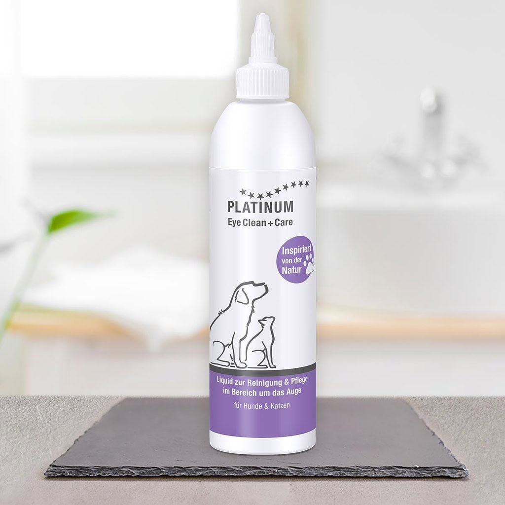 Bela steklenička PLATINUM Eye Clean + Care, Nega Oči za Pse in Mačke stoji na površini iz skrilavca. Na etiketi sta črtni risbi psa in mačke z nemškim besedilom in vijoličnim poudarkom. V ozadju je vidno zamegljeno umivalnik.