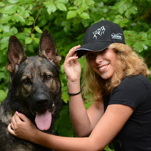 Ženska s kodrastimi svetlimi lasmi, ki nosi Ekskluzivna PLATINUM kapa s šiltom, se nasmiha ob srečnem nemškem ovčarju sredi zelenega listja - idealen navdih za ljubiteljske dodatke za kosmatince znamke PLATINUM.