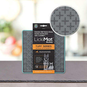 Podloga LickiMat® - TUFF BUDDY blagovne znamke PLATINUM stoji na mizi. Siva podloga, ki je zasnovana za zmanjševanje tesnobe psov, ima na površini dvignjen vzorec križa. Povečan krog prikazuje teksturo; na embalaži sta prikazana dva psa.