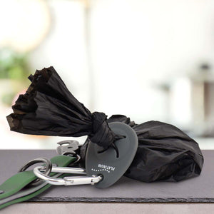 Črna vrečka za pasje iztrebke je privezana in drži jo PLATINUM Nosilec vrečk za iztrebke, pritrjena na zelen povodec s srebrno karabinko - nepogrešljiv pripomoček za vsak pasji sprehod. Predmeti so postavljeni na površino z zamegljenim notranjim ozadjem.