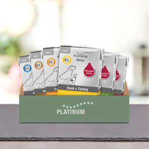 V zeleni škatli z napisom PLATINUM je prikazan Poskusni paket različnih okusov Mokre Hrane za Pse s pokončnimi paketi kakovostne pasje hrane iz sveže mesa z okusi, kot sta Duck + Turkey in 83% Fleischanteil.