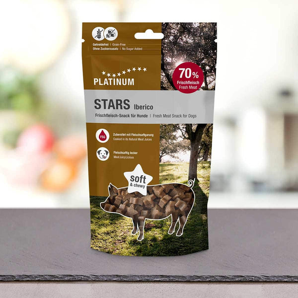 Na pultu stoji vrečka PLATINUM Iberico Stars 185 g - Nepremagljiva nagrada iz 100 % svežega mesa Iberico. Na embalaži je silhueta prašiča, napolnjena z dobrotami, v ozadju pa so vidna drevesa.