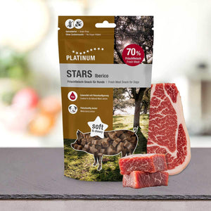 Paket PLATINUM Iberico Stars 185 g - nagrada iz 100 % svežega mesa Iberico je prikazan na pultu z zrezki in kockami mesa, pri čemer je poudarjeno "70 % svežega mesa", "mehko in žvečljivo", z ibersko svinjino in drevesom s travo v ozadju.