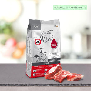 Vrečka PLATINUM Adult Beef, Suha Hrana za Pse za male pasme stoji na površini iz skrilavca poleg kosov surovega govejega mesa. Ozadje je zamegljeno, v zgornjem desnem kotu pa je prikazana zelena slovenska etiketa.