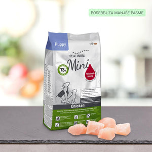 Na pultu stoji vrečka PLATINUM Puppy Chicken, Suha Hrana za Pse, pred katero so koščki surovega piščanca. Na zeleni nalepki zgoraj desno je napis "POSEBEJ ZA MANJŠE PASME". Ozadje je zamegljeno.