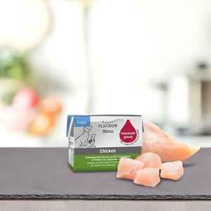 Škatla PLATINUM MENU Mini mokra hrana & DoggyCoach - Nagrada za tvojega psa stoji na pultu poleg surovega piščanca, mehko zamegljeno ozadje pa poudarja prijetno kuhinjsko okolje.